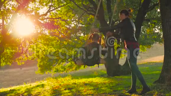 快乐的年轻妈妈和一个小女孩在日落时一起在公园里摇摆妈妈和女儿骑着秋千家人视频的预览图