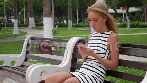 年轻漂亮的苗条女人一头金发穿着黑白连衣裙坐在长凳上用智能手机上网视频的预览图