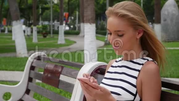 年轻漂亮的苗条女人一头金发穿着黑白连衣裙坐在长凳上用智能手机上网视频的预览图