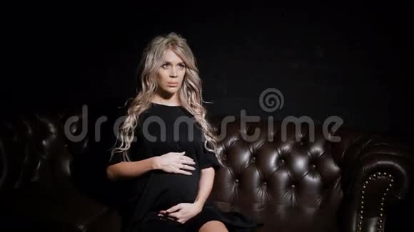 年轻的单身孕妇穿着黑色连衣裙坐在黑暗的房间里在棕色的大皮沙发上看着视频的预览图