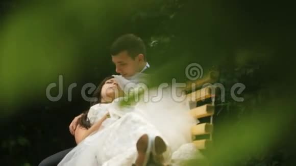 幸福的新婚夫妇在绿色阳光公园的长凳上休息新郎慈爱地抚摸他新妻子的脸视频的预览图