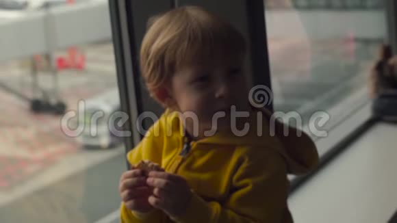 一个穿着黄色夹克的小男孩在机场的一扇大窗户前吃着饼干的慢镜头视频的预览图