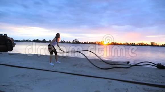 年轻的运动的金发的女人在一根厚厚的运动的绳子的帮助下进行力量练习在黎明在沙地视频的预览图