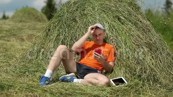 穿着橙色t恤智能手机和平板电脑的男人坐在干草堆附近视频的预览图