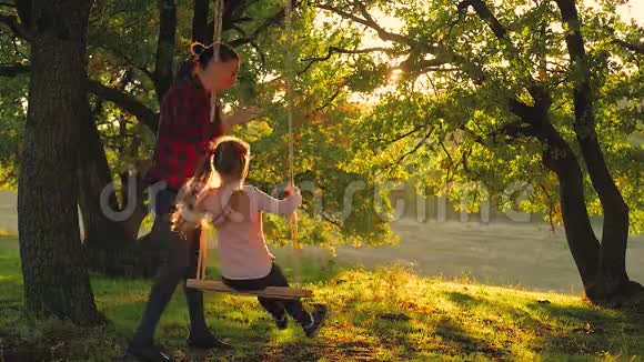 快乐的年轻妈妈和一个小女孩在日落时一起在公园里摇摆妈妈和女儿骑着秋千家人视频的预览图