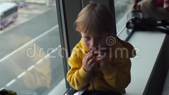 一个穿着黄色夹克的小男孩在机场的一扇大窗户前吃着饼干的慢镜头视频的预览图