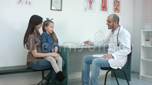 微笑男医生和害羞的小女孩坐在妈妈的腿上交谈视频的预览图