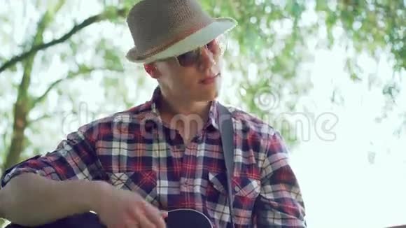 戴帽子的年轻人在阳光明媚的绿色公园里弹吉他4k视频的预览图
