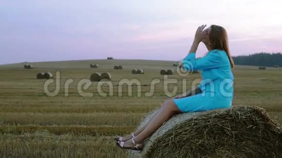 年轻漂亮的女人穿着蓝色连衣裙坐在一个大田里的干草堆上视频的预览图