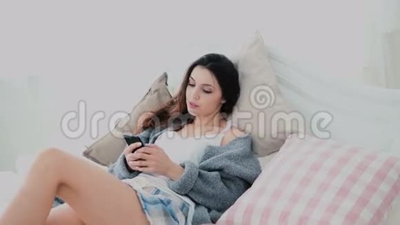 漂亮的黑发女人使用智能手机触摸屏显示在灯光室女孩躺在床上浏览互联网视频的预览图