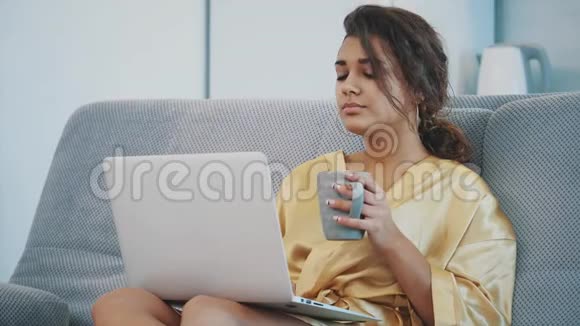 布鲁内特躺在公寓里的沙发上用笔记本电脑放松快乐的年轻女子在舒适的沙发上放松视频的预览图