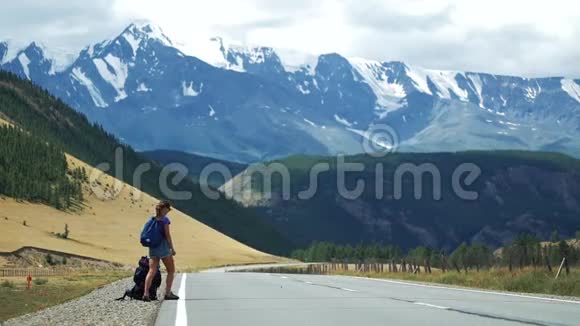 年轻的旅游妇女带着背包和太阳镜在山路上搭便车那里有雪山视频的预览图