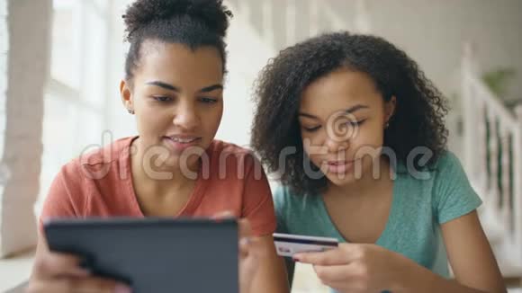 两个快乐的混血卷发女友在家用平板电脑和信用卡在网上购物视频的预览图