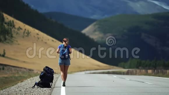 旅行者女孩带着背包和太阳镜在山路上搭便车那里有雪山视频的预览图