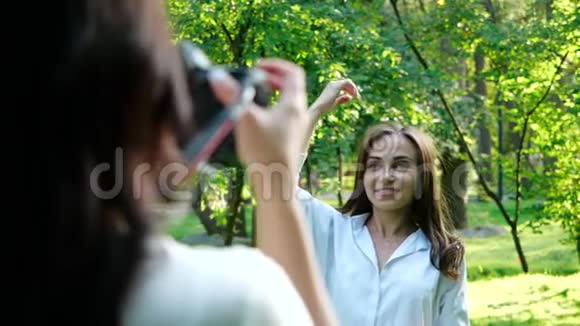一位穿着白色衬衫的漂亮女孩职业摄影师正在一个柔和的背景下拍摄一个女孩在公园里的照片视频的预览图