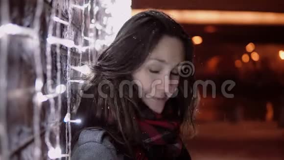 在圣诞节的夜晚一个年轻迷人的女人在飘落的雪中看着站在灯光墙附近的摄像机视频的预览图