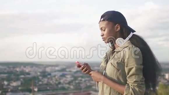 可爱的非裔美国女孩在帽子选择音乐听她的智能手机模糊的城市景观背景视频的预览图