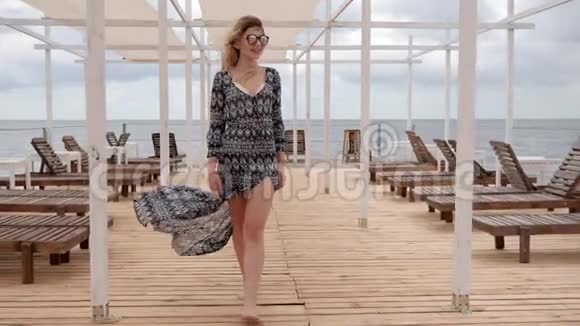 长头发的女孩在海滨开发衣服年轻的女人戴眼镜在木墩上散步昂贵的假期视频的预览图
