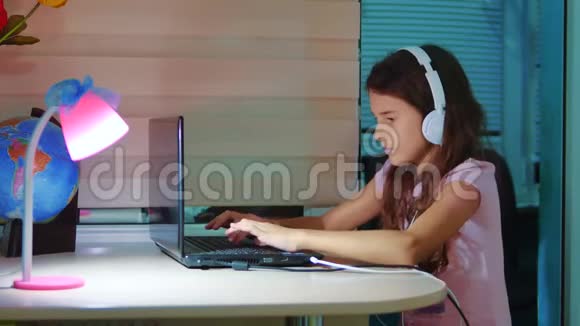 小女孩带着耳机在笔记本电脑上玩网络游戏电脑上的小女孩在社交媒体上留言视频的预览图
