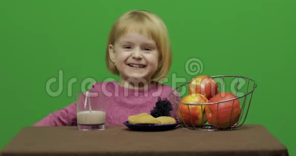 女孩坐在桌子旁吃巧克力饼干和饮料可可快乐的三岁女孩可爱的女孩微笑视频的预览图