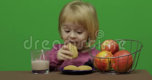 女孩坐在桌子旁吃巧克力饼干和饮料可可快乐的三岁女孩可爱的女孩微笑视频的预览图