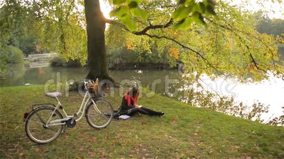 美丽的白种人微笑的女孩骑着自行车坐在公园里学生在湖边的笔记本上写着温暖视频的预览图