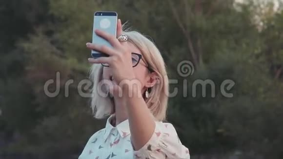 在湖边的城市公园里年轻的美女带着智能手机自拍视频的预览图