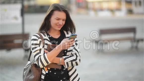 美丽的年轻女子在城市街道上使用智能手机学生女孩拿着电话和笔记本老城视频的预览图