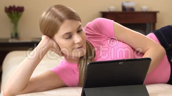 美丽的年轻女子带着一块平板电脑轻松地躺在沙发上浏览互联网和微笑视频的预览图