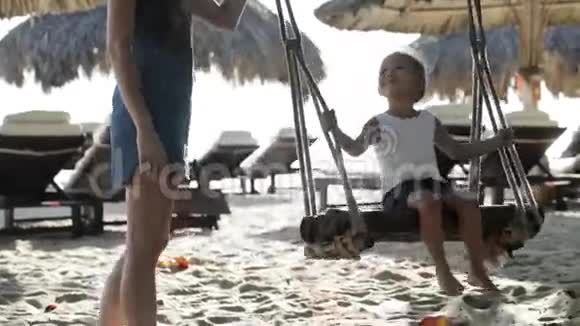 年轻的母亲推着可爱的小女儿在沙滩上荡秋千视频的预览图