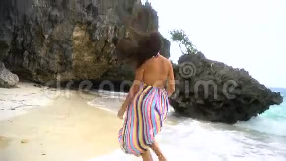 年轻的黑发女孩穿着一件鲜艳的连衣裙沿着白色的海滩奔跑微笑着看着相机着头发视频的预览图
