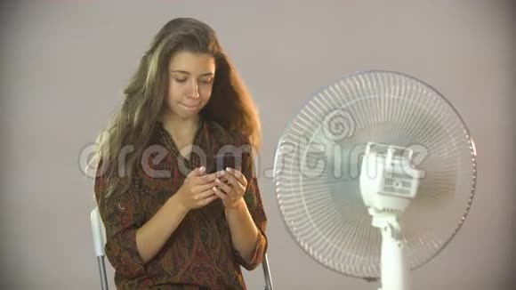 漂亮的女孩坐在风扇附近的椅子上在手机上写短信视频的预览图