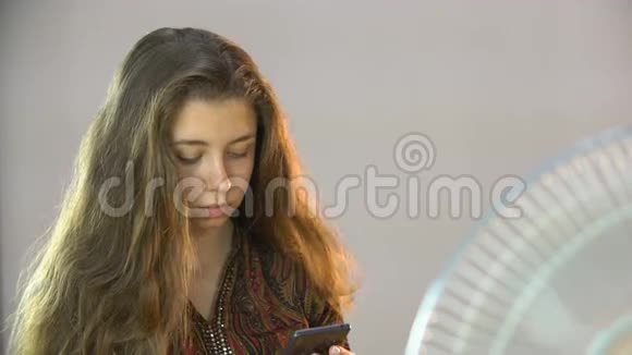 美丽的女孩坐在风扇附近的椅子上在手机上写短信视频的预览图