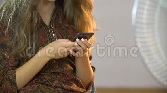 美丽的女孩坐在风扇附近的椅子上在手机上写短信视频的预览图