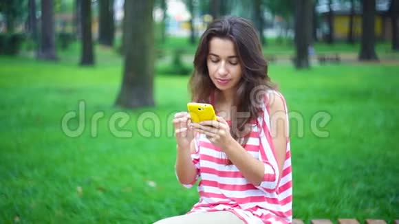 年轻女子坐在夏季公园的长椅上手里拿着智能手机视频的预览图