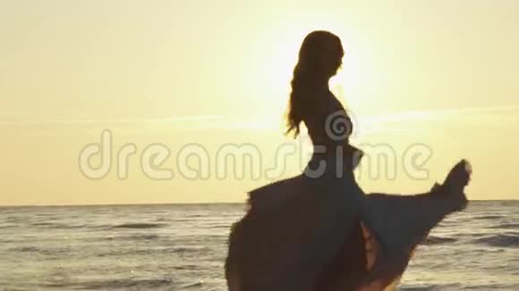 年轻迷人的女孩穿着淡粉色的长裙在金色的晚霞照耀下的沙滩上旋转视频的预览图