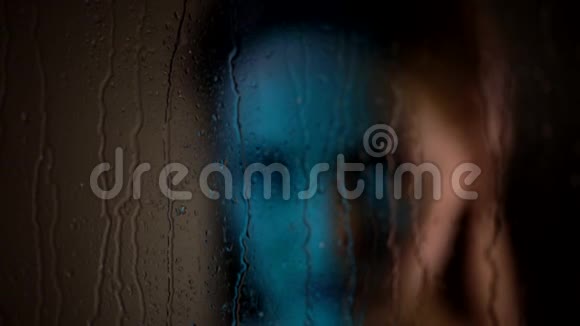近距离拍摄了一个悲伤梦幻迷人的白种人女人站在蒸汽窗户玻璃旁边看着雨滴飘落视频的预览图