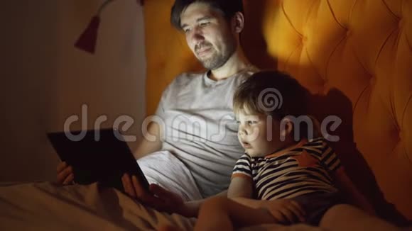 年轻的父亲和他的小儿子晚上躺在家里用平板电脑看卡通电影视频的预览图