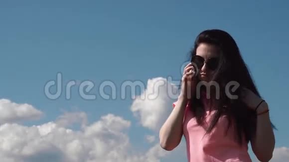 年轻迷人的黑发女孩戴着太阳镜在蓝天的阴云下通过电话交谈视频的预览图