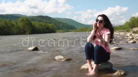 年轻风趣有点生气的黑发女人坐在河边用智能手机和别人说话视频的预览图