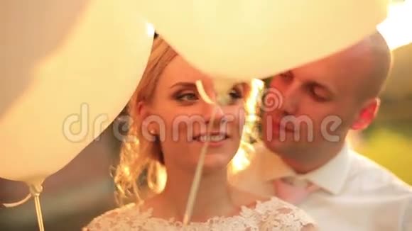 快乐的年轻新婚夫妇在公园日落时带着气球英俊的新郎温柔地亲吻他美丽的金发新娘视频的预览图