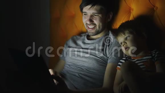 年轻的父亲和他的小儿子晚上躺在床上用平板电脑看动画片聊天视频的预览图