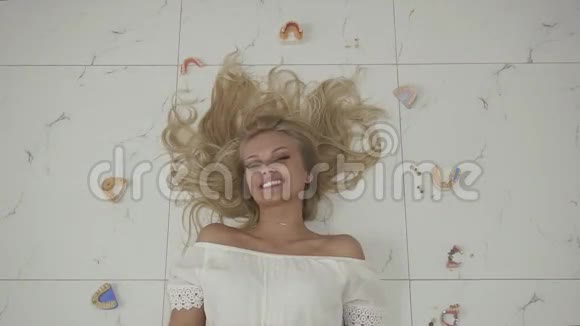美丽的金发女孩微笑着躺在地板上的相机假牙掉在周围牙齿健康概念视频的预览图