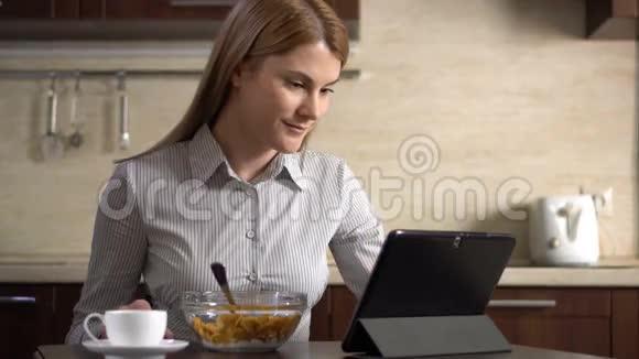 美丽的年轻迷人的女商人早餐吃玉米片并使用她的平板电脑视频的预览图