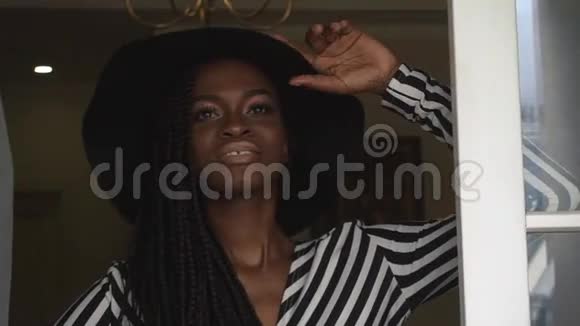 一位戴黑帽子穿着条纹连衣裙的优雅时尚的非裔美国妇女在豪华公寓里摆姿势的特写镜头视频的预览图