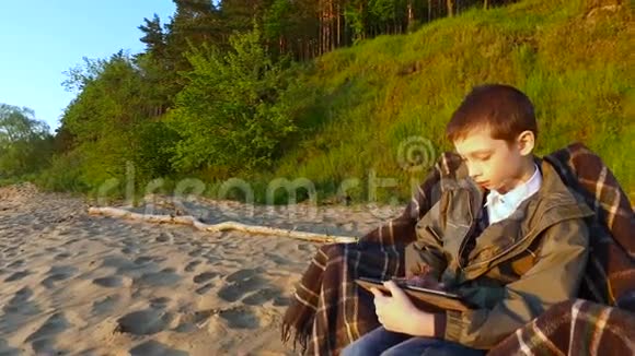少年男孩坐在沙地上的椅子上十几岁的男孩热衷于在平板电脑上玩电脑游戏视频的预览图