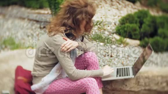 一个拿着笔记本电脑的女人在一个美丽的绿色公园的岩石上放松一位常年在植物园工作的年轻女子视频的预览图