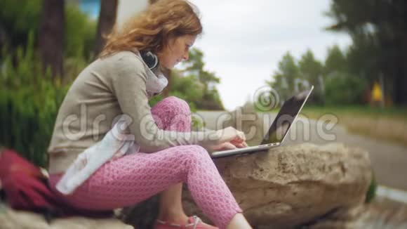 一个拿着笔记本电脑的女人在一个美丽的绿色公园的岩石上放松一位常年在植物园工作的年轻女子视频的预览图