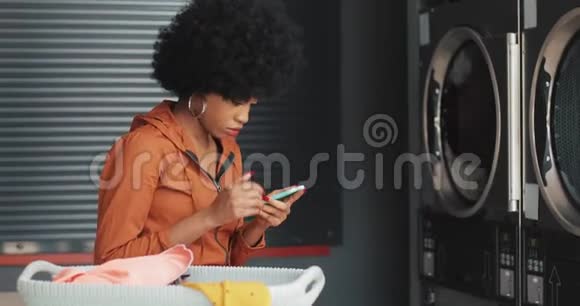在洗衣店用智能手机微笑的年轻女人自助公共洗衣店视频的预览图