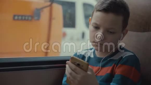 一个小男孩乘公共汽车穿过城市的特写镜头他在智能手机上使用社交网络倾听视频的预览图